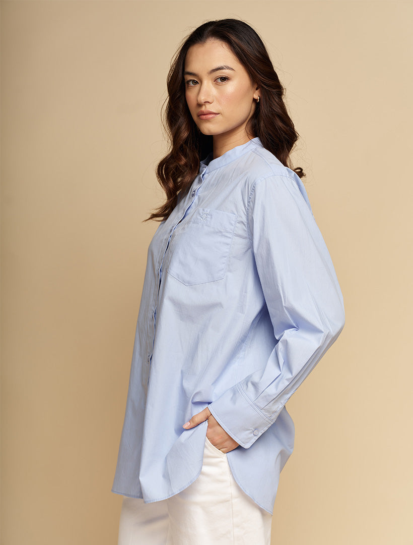 A woman wearing a light blue long-sleeve button-up shirt and white pants, standing against a solid beige studio background.