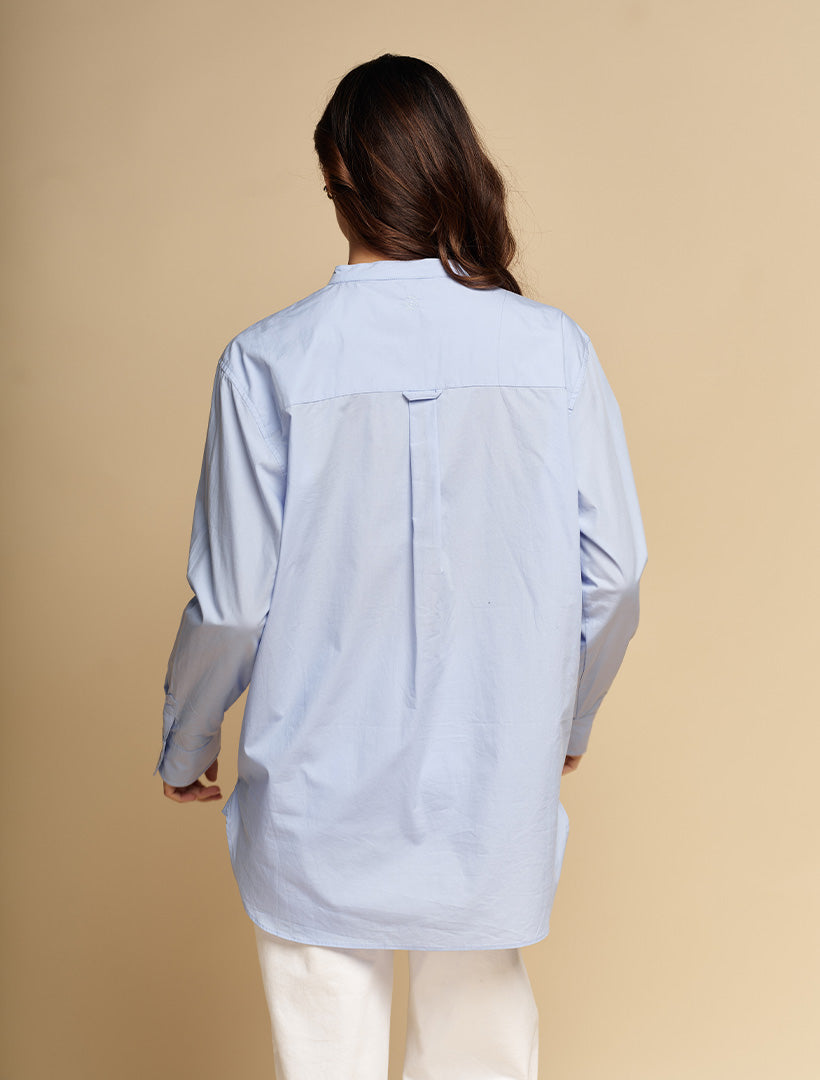 A woman viewed from behind wearing a light blue oversized button-up shirt with contrasting light gray sleeves and white pants, set against a solid beige studio background.