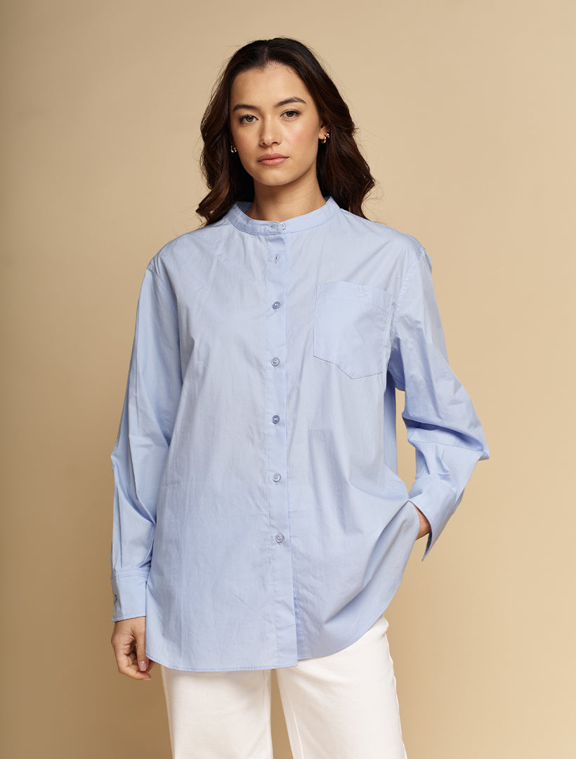 A woman modeling a light blue long-sleeve button-up shirt with a band collar and a chest pocket, paired with white pants, against a solid beige studio background.