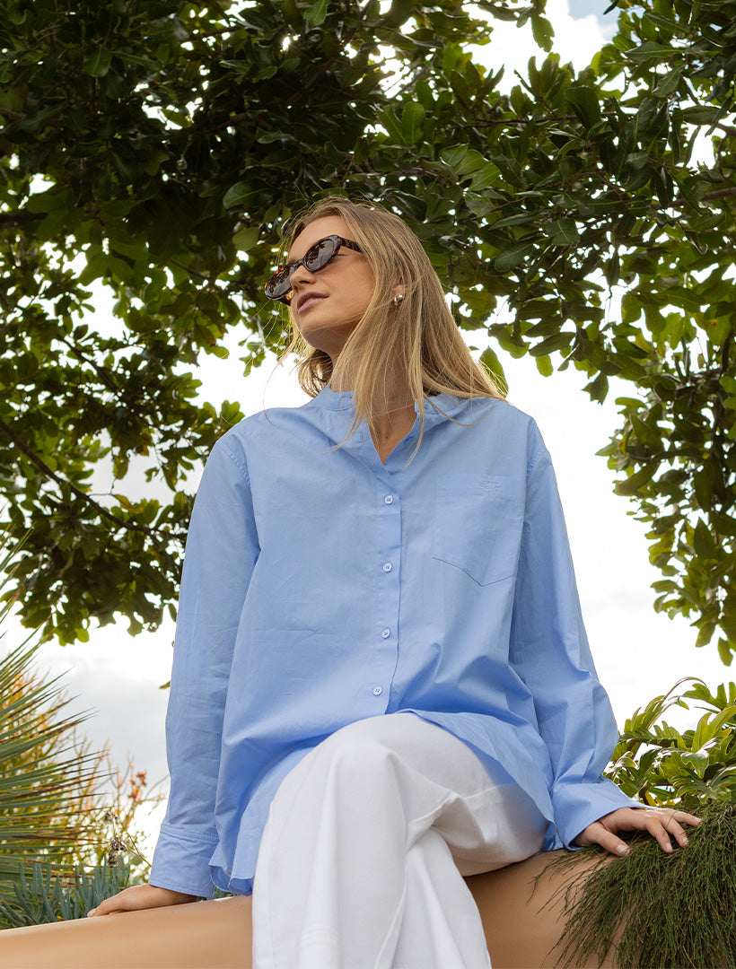 A woman wearing a light blue long-sleeve button-up shirt and white pants sits outdoors under a leafy tree, wearing dark sunglasses.