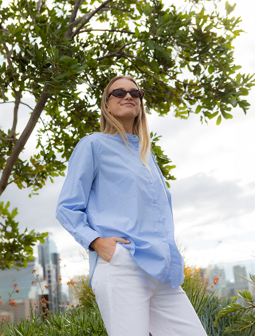 A woman wearing a light blue long-sleeve button-up shirt and white pants stands outdoors under a tree with a cloudy sky and distant city skyline in the background.